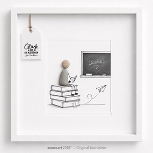 May include: A white framed wall art piece featuring a pebble art figure sitting on a stack of illustrated books. The figure holds a small book, facing a chalkboard with the text Danke! and a heart. A paper airplane and a hanging tag with text are included.