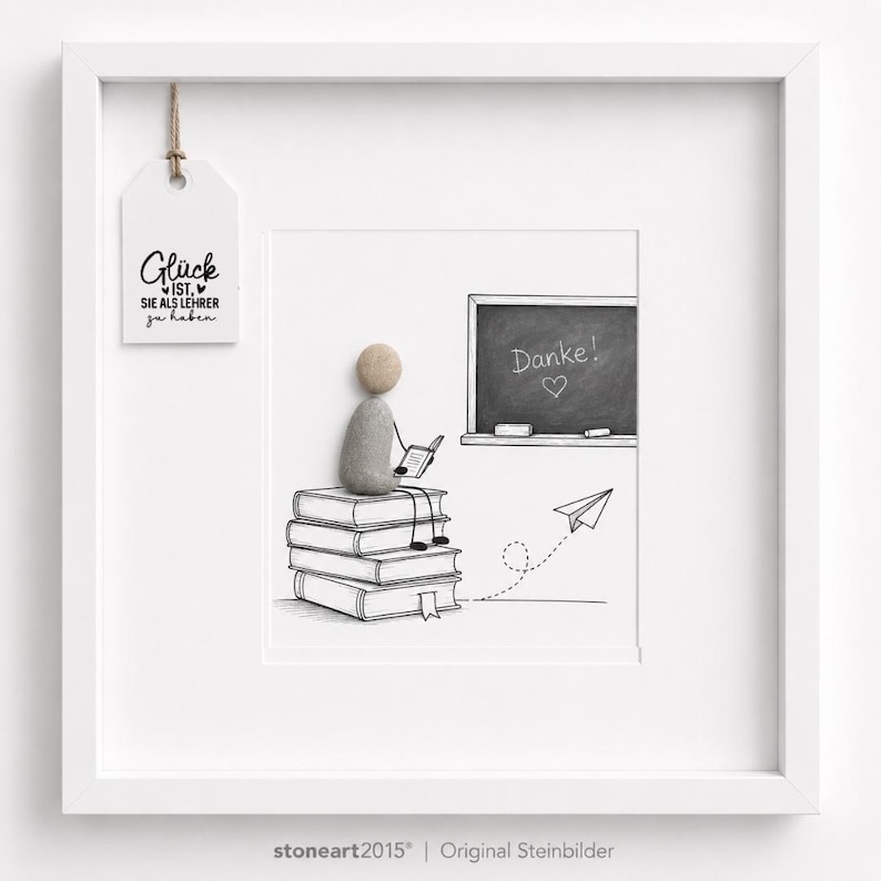 May include: A white framed wall art piece featuring a pebble art figure sitting on a stack of illustrated books. The figure holds a small book, facing a chalkboard with the text Danke! and a heart. A paper airplane and a hanging tag with text are included.