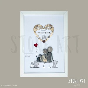 May include: Framed artwork featuring a family depicted with stones on a bench, with a red balloon. Above, a wooden heart with text in German reads "Holzernen Hochzeit". The frame is white.