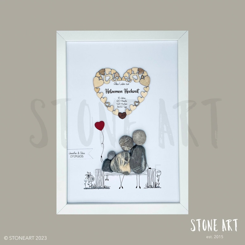 May include: Framed artwork featuring a family depicted with stones on a bench, with a red balloon. Above, a wooden heart with text in German reads "Holzernen Hochzeit". The frame is white.