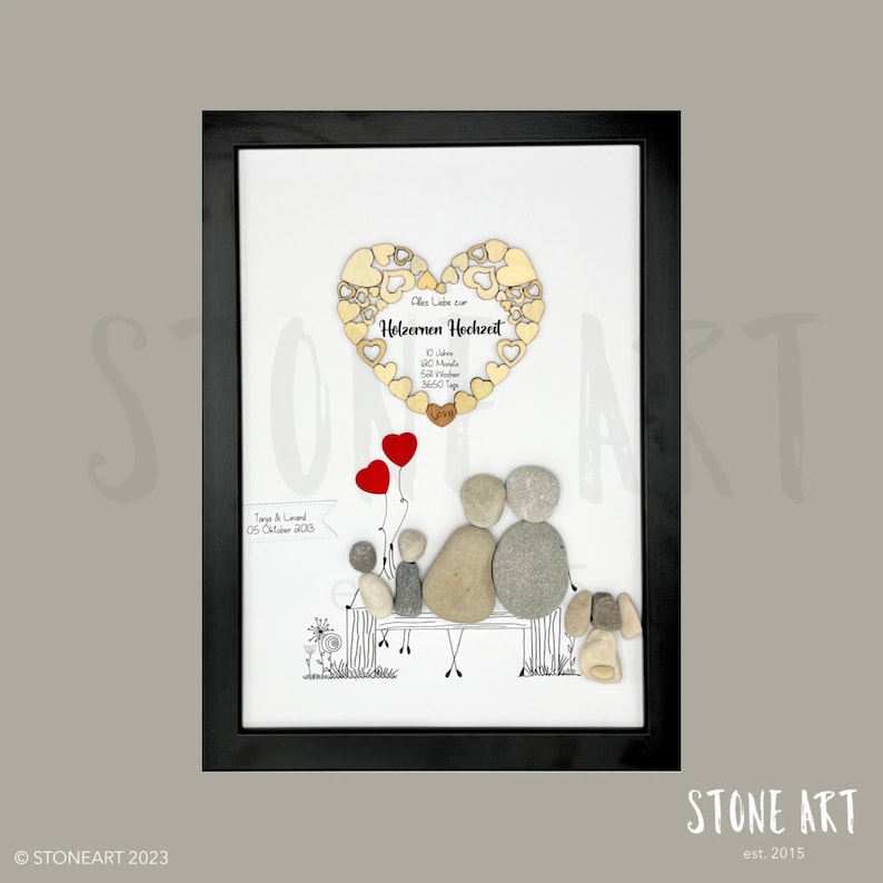 May include: Framed artwork featuring a family depicted with stones on a bench, with red heart balloons. A wooden heart with text in German is at the top. The artwork is in a black frame.