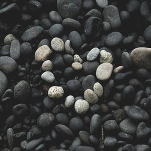 May include: A collection of dark gray and light tan river rocks arranged in a heart shape. The heart is surrounded by a dense field of similar stones, creating a textured, natural pattern. The image has a moody, monochromatic aesthetic.