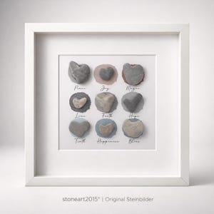 Può includere: Opera d'arte incorniciata con nove pietre a forma di cuore, ognuna con una parola come "Pace" o "Gioia". Le pietre sono disposte in una griglia, su uno sfondo bianco con cerchi dai colori tenui. La cornice è bianca.
