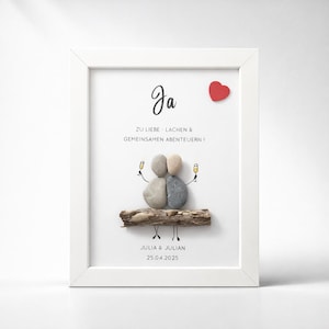 May include: A white framed wall art piece featuring pebble art of two figures sitting on a piece of driftwood, holding champagne glasses. The text reads Ja, ZU LIEBE - LACHEN & GEMEINSAMEN ABENTEUERN!, JULIA & JULIAN, and 25.04.2025 with a red heart.