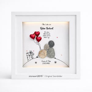 Può includere: Un'opera d'arte incorniciata con un bordo bianco. L'opera presenta una scena di arte di ciottoli di una coppia con palloncini a forma di cuore rossi. Il testo include "Rubin Hochzeit" e i nomi "Karina & Peter" con una data. Il testo in basso recita "stoneart2015 | Original Steinbilder".