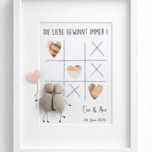 Pode incluir: Obra de arte emoldurada com o texto "DIE LIEBE GEWINNT IMMER!" e um jogo da velha com peças em forma de coração. Apresenta um casal de arte em pedra segurando um balão de coração rosa. Os nomes "Eva & Max" e a data "06. Juni 2026" também estão incluídos.