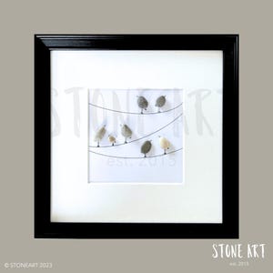 Puede incluir: Una obra de arte de guijarros enmarcada que representa a un grupo de pájaros grises y blancos posados en un alambre. El alambre está tendido entre dos postes y los pájaros miran en diferentes direcciones. El marco es negro y el paspartú es blanco. El texto "est. 2015" está impreso en negro en la parte inferior del paspartú.