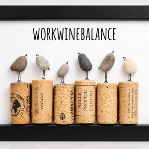 May include: A framed wall art piece featuring six small birds made of smooth grey and beige pebbles perched on top of six vertical wine corks. The text WORKWINEBALANCE is printed in black sans-serif font above the birds on a white background.