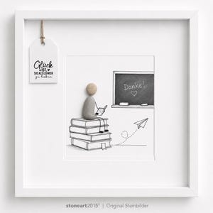 May include: A white framed wall art piece featuring a pebble art figure sitting on a stack of illustrated books. The figure holds a small book, facing a chalkboard with the text Danke! and a heart. A paper airplane and a hanging tag with text are included.