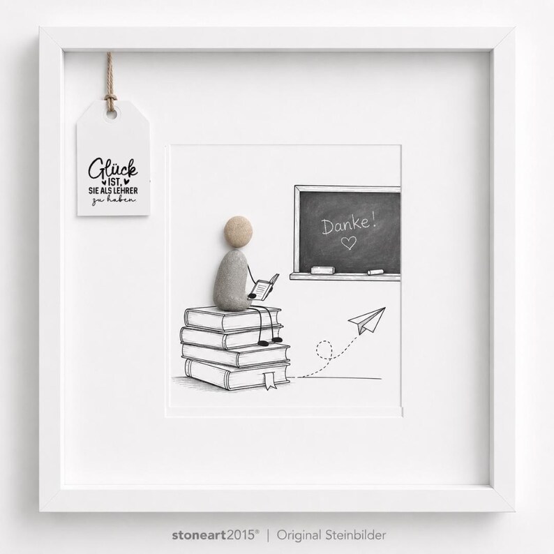 May include: A white framed wall art piece featuring a pebble art figure sitting on a stack of illustrated books. The figure holds a small book, facing a chalkboard with the text Danke! and a heart. A paper airplane and a hanging tag with text are included.