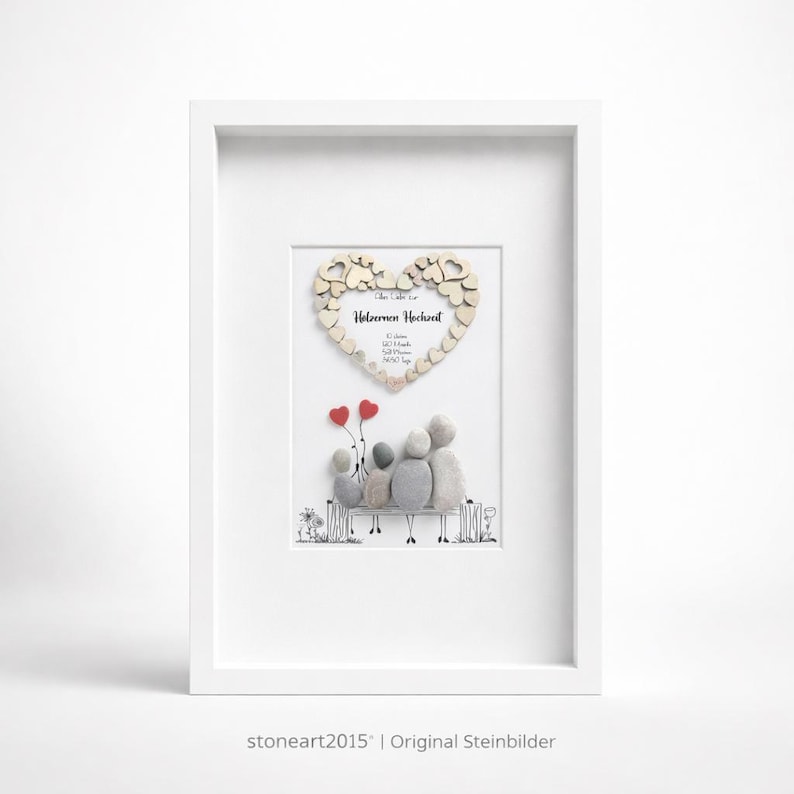 May include: Framed artwork featuring a family of stone figures seated on a bench, with red heart balloons. A wooden heart with text is above the family. The artwork is in a white frame.