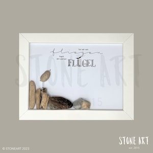 Könnte beinhalten: Ein weiß gerahmtes Bild mit weißem Hintergrund und schwarzem Text, der "Nur wer vom traumt, dem wachsen FLUGEL" lautet. Der Text ist in einer kursiven Schrift geschrieben. Am unteren Rand des Bildes befindet sich ein kleiner Vogel aus Steinen auf einem Haufen Treibholz. Das Bild ist in einem weißen Rahmen gerahmt.