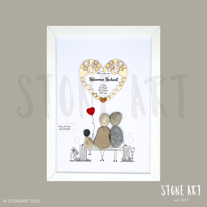 May include: Framed artwork featuring a pebble art family on a bench, holding a red balloon. Above, a wooden heart with text in German. The artwork is in a white frame, with the text "STONE ART" and the copyright notice at the bottom.