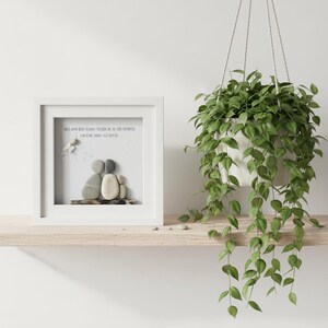 May include: A framed pebble art piece depicting a family of three, with a white frame and a light-colored wooden shelf. The art includes a quote in German. A hanging plant with green leaves is to the right.