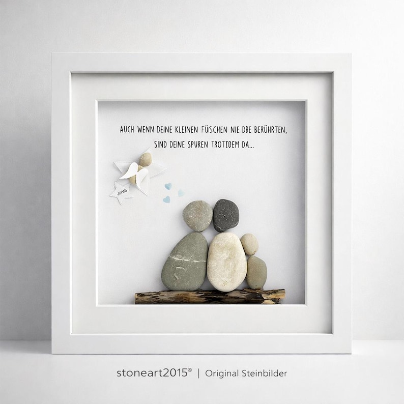 May include: A framed artwork featuring a family depicted with smooth stones on a piece of wood. The artwork includes an angel and text in German. The frame is white, and the background is also white.