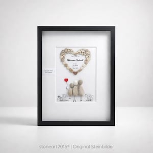 May include: Framed artwork featuring a heart-shaped design made of small, light-colored stones. Below the heart, three figures sit on a bench, with a red heart-shaped balloon. The artwork is in a black frame.
