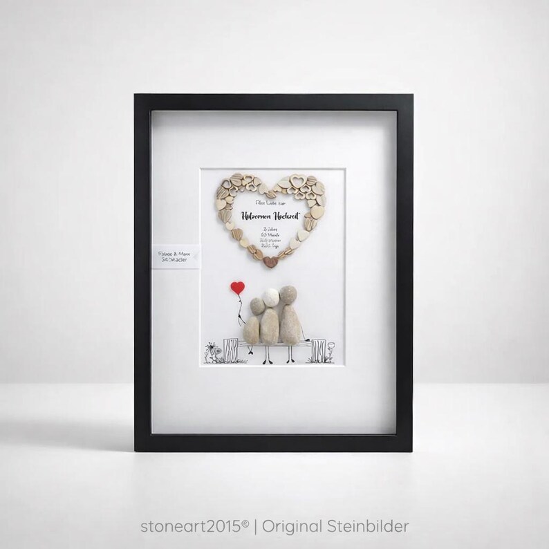 May include: Framed artwork featuring a heart-shaped design made of small, light-colored stones. Below the heart, three figures sit on a bench, with a red heart-shaped balloon. The artwork is in a black frame.
