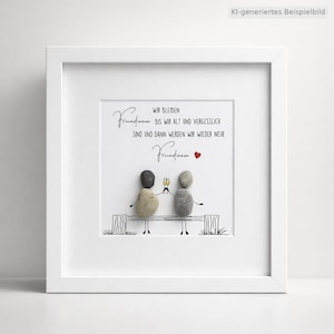 May include: A square white frame containing a pebble art piece. Two figures made of grey and beige stones sit on a drawn bench, holding small yellow glasses. The text reads: Wir bleiben Freundinnen bis wir alt und vergesslich sind und dann werden wir wieder neue Freundinnen.