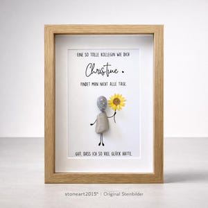 Op de afbeelding: Ingelijst kunstwerk met een licht houten frame. De kunst toont een kiezelsteenfiguur met een gele zonnebloem. De Duitse tekst luidt: "EINE SO TOLLE KOLLEGIN WIE DICH Christine. FINDET MAN NICHT ALLE TAGE. GUT, DASS ICH SO VIEL GLÜCK HATTE."