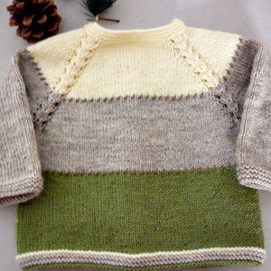 May include: A knitted baby sweater with a striped pattern in beige, light brown, and green. The sweater has a white crocheted trim around the neckline and sleeves.