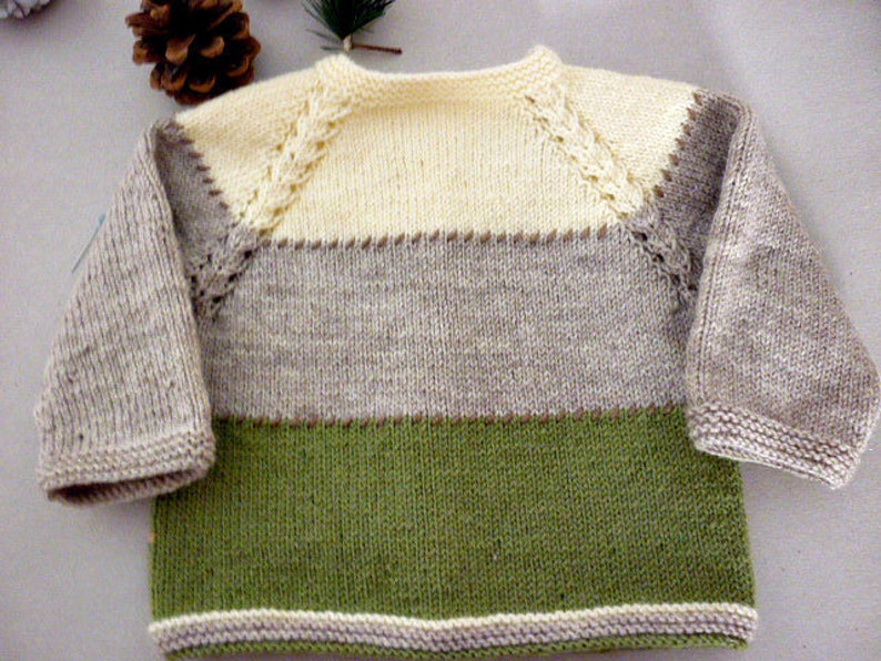 May include: A knitted baby sweater with a striped pattern in beige, light brown, and green. The sweater has a white crocheted trim around the neckline and sleeves.