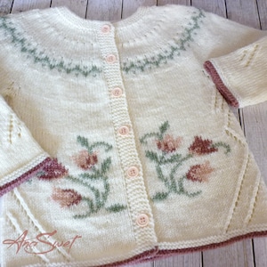 May include: A cream-colored knitted cardigan with a floral design and a button-up front. The cardigan has a green and white pattern around the neckline and a brown trim around the sleeves and bottom.