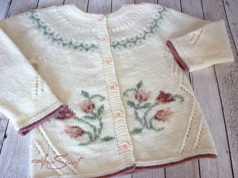 May include: A cream-colored knitted cardigan with a floral design and a button-up front. The cardigan has a green and white pattern around the neckline and a brown trim around the sleeves and bottom.
