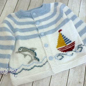 May include: A white and blue striped knitted baby cardigan with a dolphin and sailboat design. The cardigan has a button closure and is made of soft yarn.