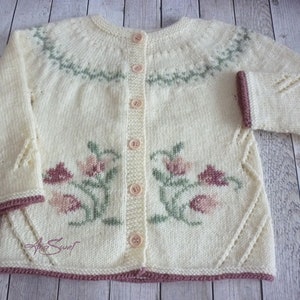May include: A cream-colored knitted cardigan with a floral design in pink, green, and brown. The cardigan has a button closure and a ribbed trim.
