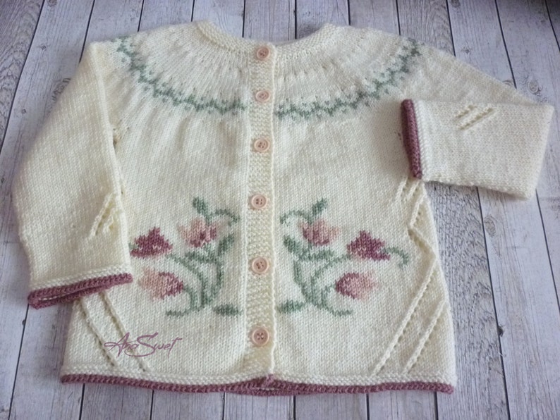 May include: A cream-colored knitted cardigan with a floral design in pink, green, and brown. The cardigan has a button closure and a ribbed trim.