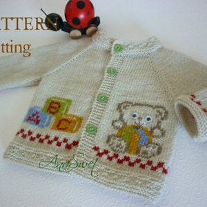 Op de afbeelding: Een gebreide baby-trui in crèmekleur met een teddybeer- en alfabetblokkenontwerp. De trui heeft rode en groene bies en groene knopen.