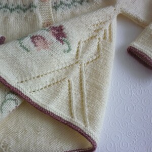 May include: A white knitted cardigan with a brown trim and floral embroidery. The cardigan has a button closure and a delicate lace pattern on the sleeves.