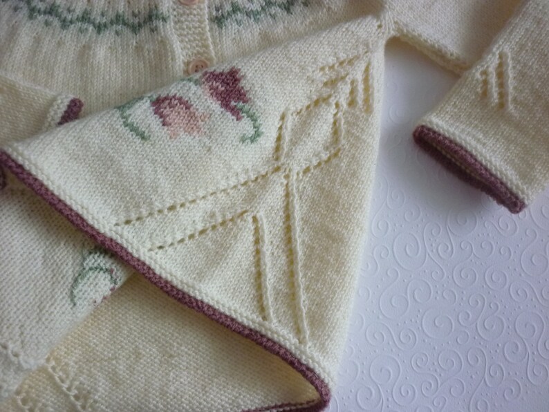 May include: A white knitted cardigan with a brown trim and floral embroidery. The cardigan has a button closure and a delicate lace pattern on the sleeves.