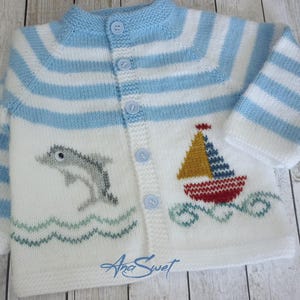 May include: A white knitted baby cardigan with blue and white stripes. The cardigan has a dolphin and a sailboat embroidered on the front. The cardigan has a button closure.
