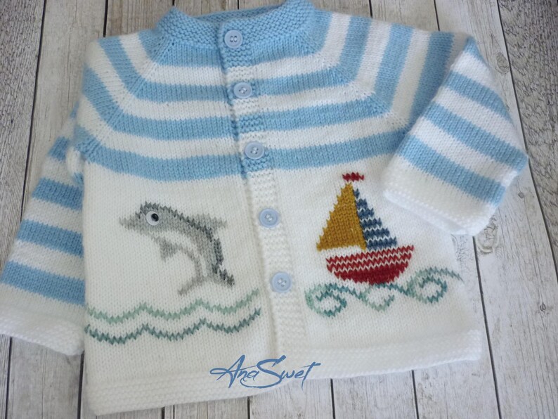 May include: A white knitted baby cardigan with blue and white stripes. The cardigan has a dolphin and a sailboat embroidered on the front. The cardigan has a button closure.