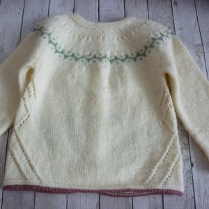 May include: A cream-colored knitted sweater with a green and white geometric pattern around the neckline. The sweater has a pink trim around the bottom and sleeves.