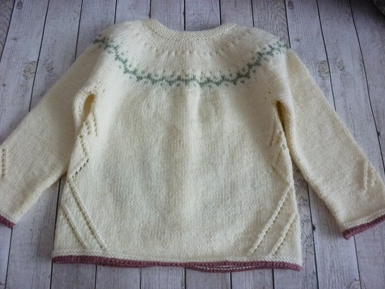 May include: A cream-colored knitted sweater with a green and white geometric pattern around the neckline. The sweater has a pink trim around the bottom and sleeves.
