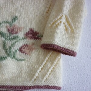 May include: A white knitted cardigan with a floral design in shades of pink and green. The cardigan has a button closure and a scalloped edge.