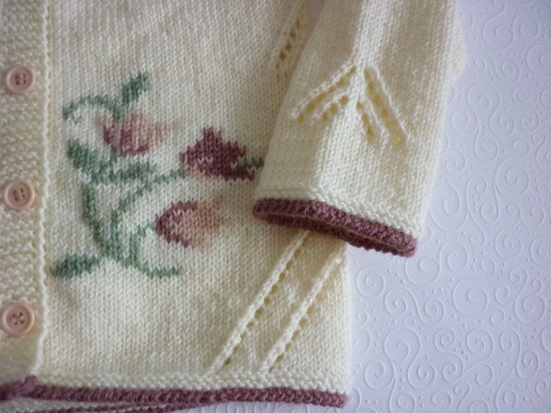 May include: A white knitted cardigan with a floral design in shades of pink and green. The cardigan has a button closure and a scalloped edge.