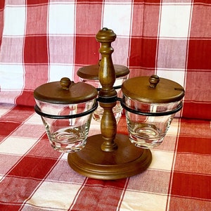 May include: A wooden condiment server with three glass jars, each with a wooden lid. The jars are held in place by a dark metal ring. The server is on a red and white checkered tablecloth.
