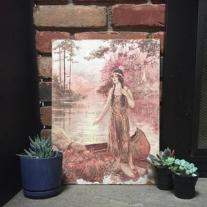 May include: Vintage print of a Native American woman in a canoe with the text "Little, Strong the Tryst". The print is framed and hanging on a brick wall.