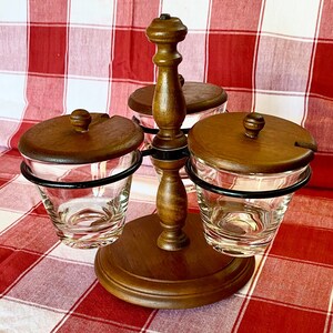 K&ouml;nnte beinhalten: Ein h&ouml;lzerner Gew&uuml;rzst&auml;nder mit drei durchsichtigen Gl&auml;sern, jedes mit einem Holzdeckel und einer kleinen L&ouml;ffelkerbe. Die Gl&auml;ser werden von schwarzen Metallringen gehalten. Der St&auml;nder hat eine dunkle Holzoberfl&auml;che und steht auf einem runden Sockel.