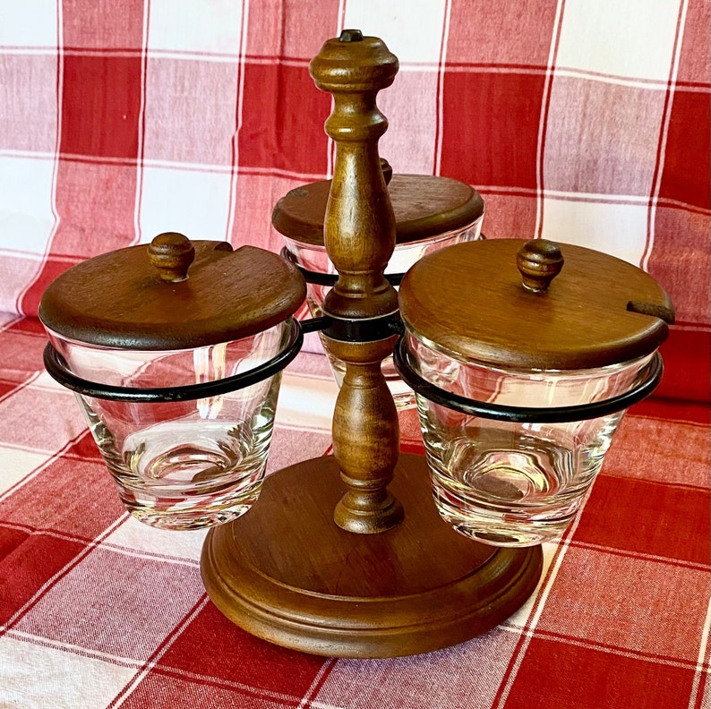 K&ouml;nnte beinhalten: Ein h&ouml;lzerner Gew&uuml;rzst&auml;nder mit drei durchsichtigen Gl&auml;sern, jedes mit einem Holzdeckel und einer kleinen L&ouml;ffelkerbe. Die Gl&auml;ser werden von schwarzen Metallringen gehalten. Der St&auml;nder hat eine dunkle Holzoberfl&auml;che und steht auf einem runden Sockel.