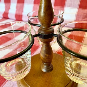 K&ouml;nnte beinhalten: Ein h&ouml;lzerner Schnapsglashalter mit drei durchsichtigen Schnapsgl&auml;sern. Der Halter hat eine dunkelbraune Oberfl&auml;che und einen kreisf&ouml;rmigen Sockel. Die Schnapsgl&auml;ser werden von schwarzen Metallringen gehalten.