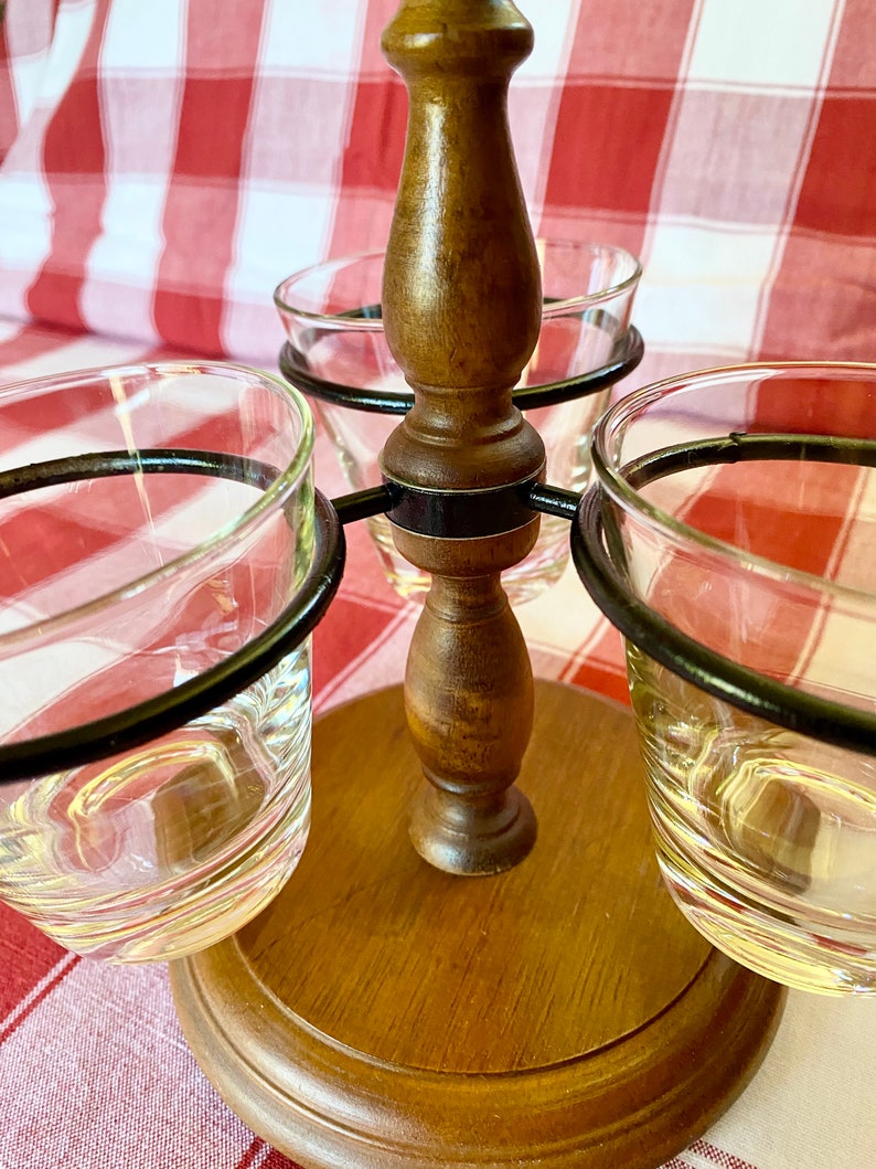 K&ouml;nnte beinhalten: Ein h&ouml;lzerner Schnapsglashalter mit drei durchsichtigen Schnapsgl&auml;sern. Der Halter hat eine dunkelbraune Oberfl&auml;che und einen kreisf&ouml;rmigen Sockel. Die Schnapsgl&auml;ser werden von schwarzen Metallringen gehalten.