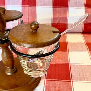 K&ouml;nnte beinhalten: Ein Vintage-Gew&uuml;rzset mit durchsichtigen Gl&auml;sern, Holzdeckeln und einem kleinen L&ouml;ffel. Die Gl&auml;ser werden von einem dunklen Metallring gehalten und auf einem Holzst&auml;nder montiert. Der Hintergrund ist eine rot-wei&szlig; karierte Tischdecke.