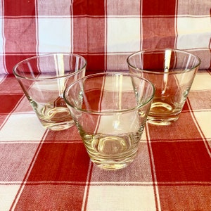K&ouml;nnte beinhalten: Drei klare, konische Trinkgl&auml;ser. Die Gl&auml;ser bestehen aus transparentem Glas und haben einen leicht ausgestellten Rand. Sie sind auf einer rot-wei&szlig; karierten Tischdecke angeordnet.