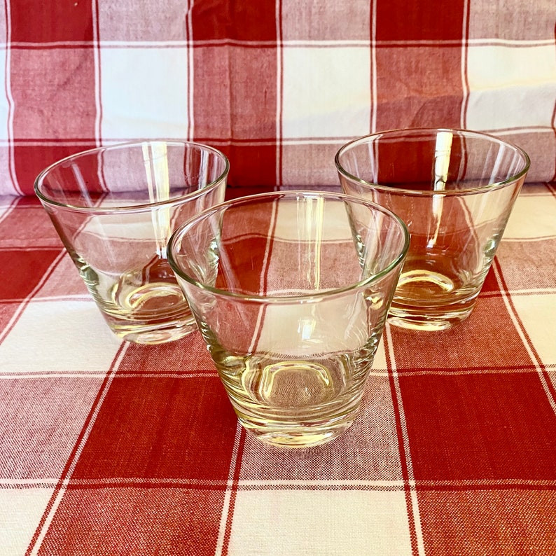 K&ouml;nnte beinhalten: Drei klare, konische Trinkgl&auml;ser. Die Gl&auml;ser bestehen aus transparentem Glas und haben einen leicht ausgestellten Rand. Sie sind auf einer rot-wei&szlig; karierten Tischdecke angeordnet.