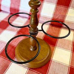 K&ouml;nnte beinhalten: Ein Weinhalter aus Holz mit rundem Sockel und Mittels&auml;ule. Drei schwarze Metallringe erstrecken sich von der S&auml;ule, um Weinflaschen zu halten. Der Halter ist aus hellem und dunklem braunem Holz gefertigt.