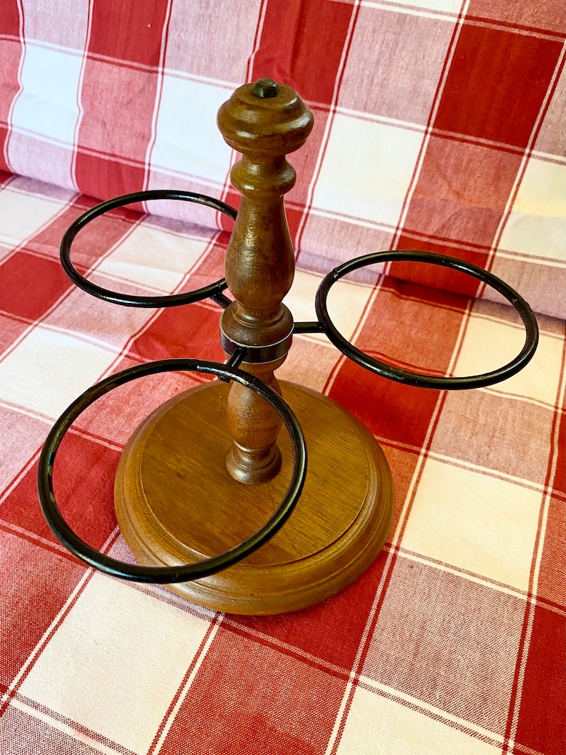 K&ouml;nnte beinhalten: Ein Weinhalter aus Holz mit rundem Sockel und Mittels&auml;ule. Drei schwarze Metallringe erstrecken sich von der S&auml;ule, um Weinflaschen zu halten. Der Halter ist aus hellem und dunklem braunem Holz gefertigt.